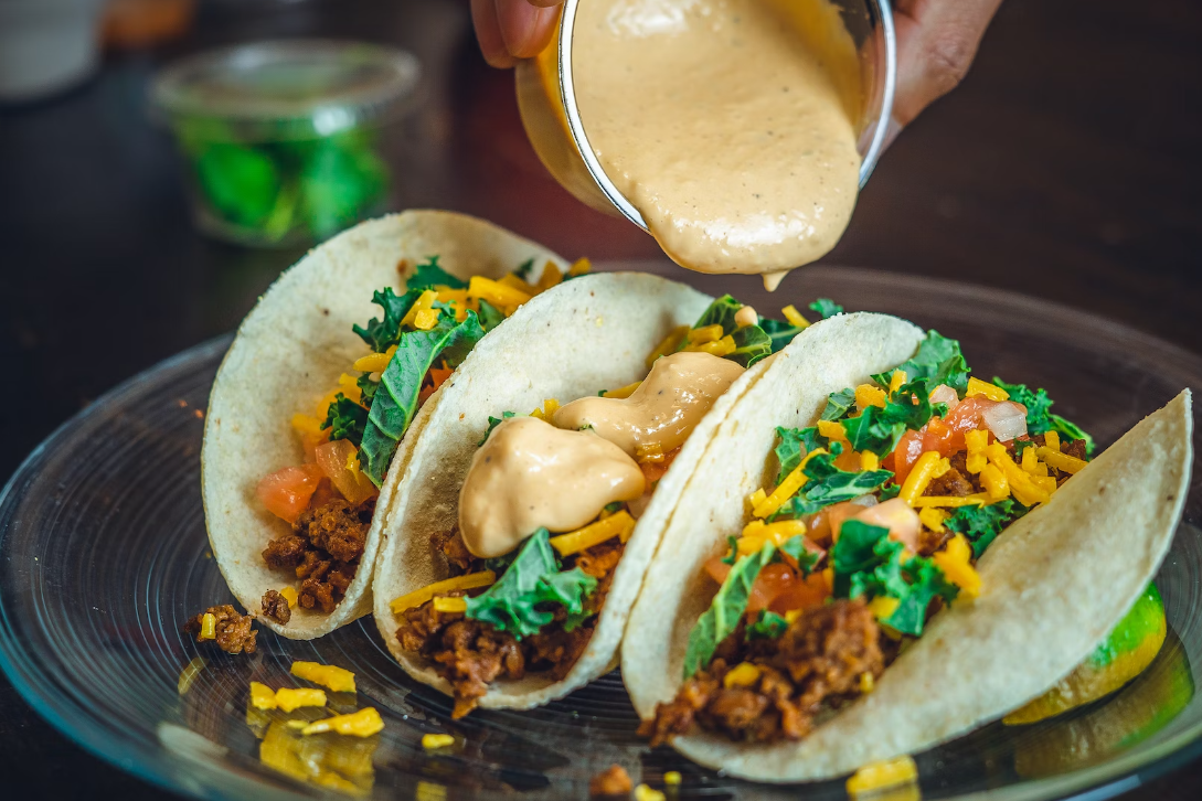 A plate of tacos with cheese, lettuce, and sauce being poured on top.
