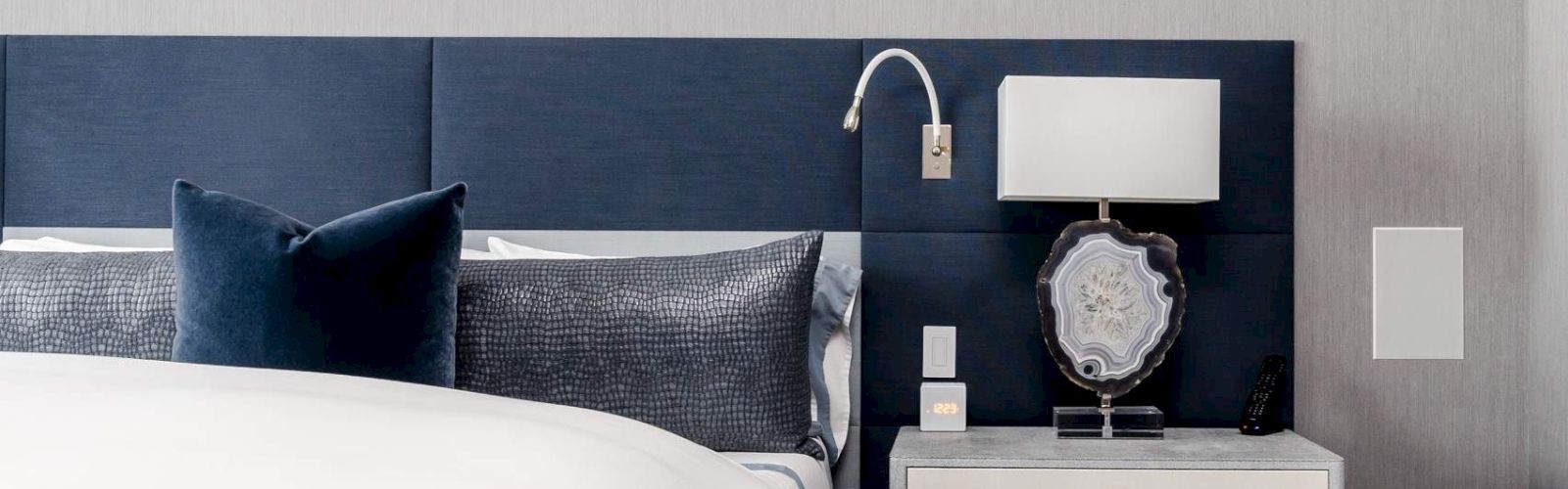 A close-up of a hotel bed with a blue headboard, white bedding, and a nightstand with a lamp.