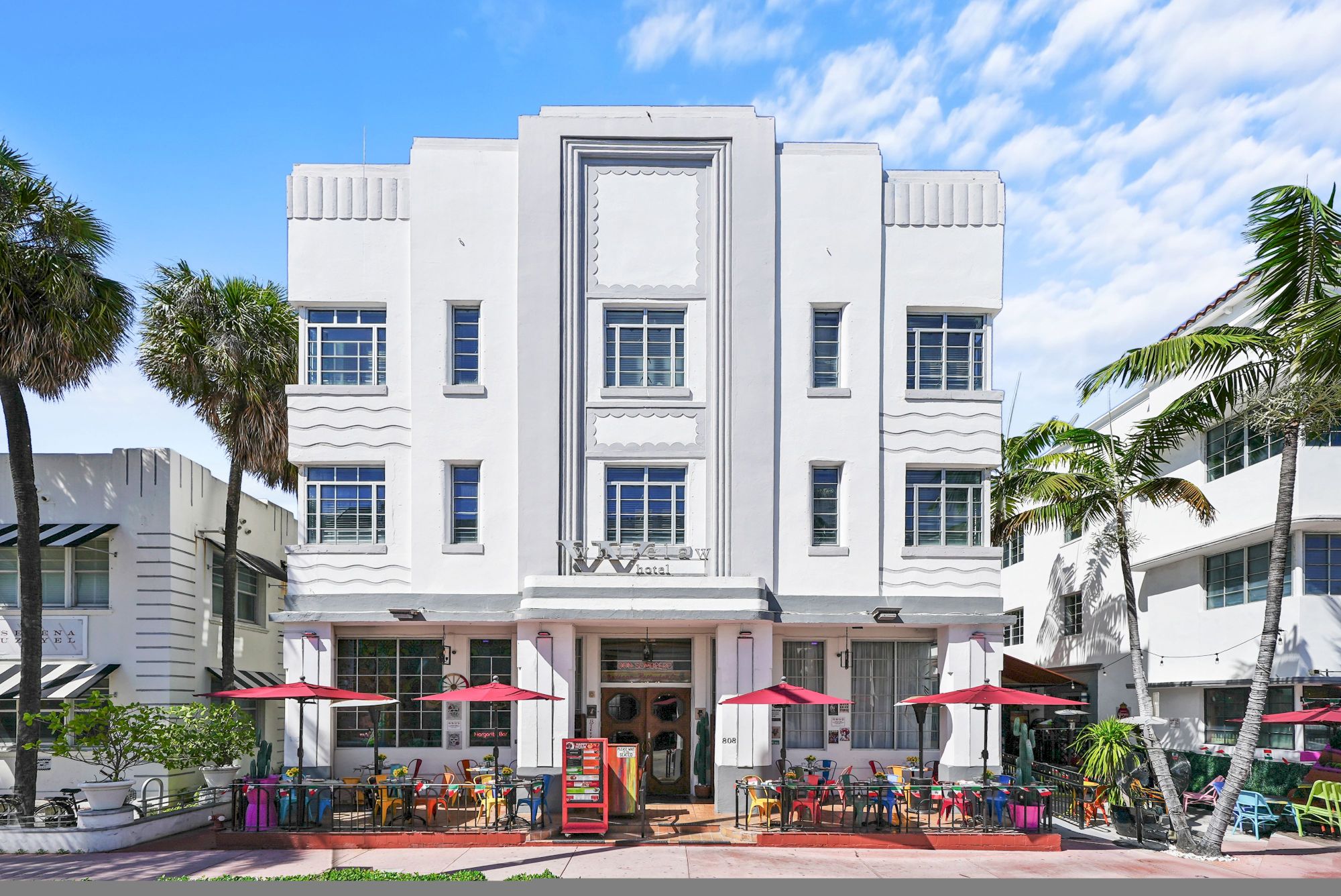 A stylish white Art Deco-style hotel with a central entrance, outdoor seating under red umbrellas, and palm trees lining the sidewalk.