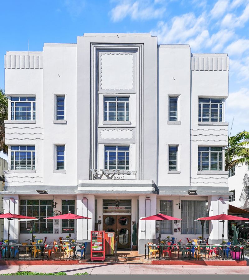 A white, Art Deco-style hotel facade with a patio area featuring colorful chairs and red umbrellas.