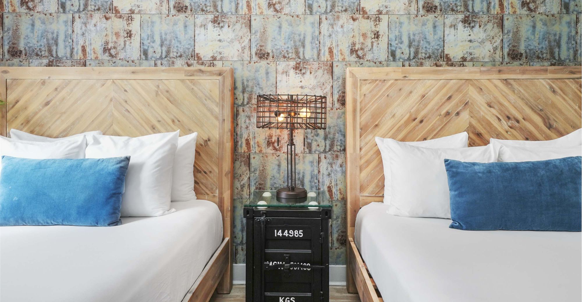 Two neatly made twin beds with wooden headboards, white linens, blue pillows, and a shared vintage nightstand between them.