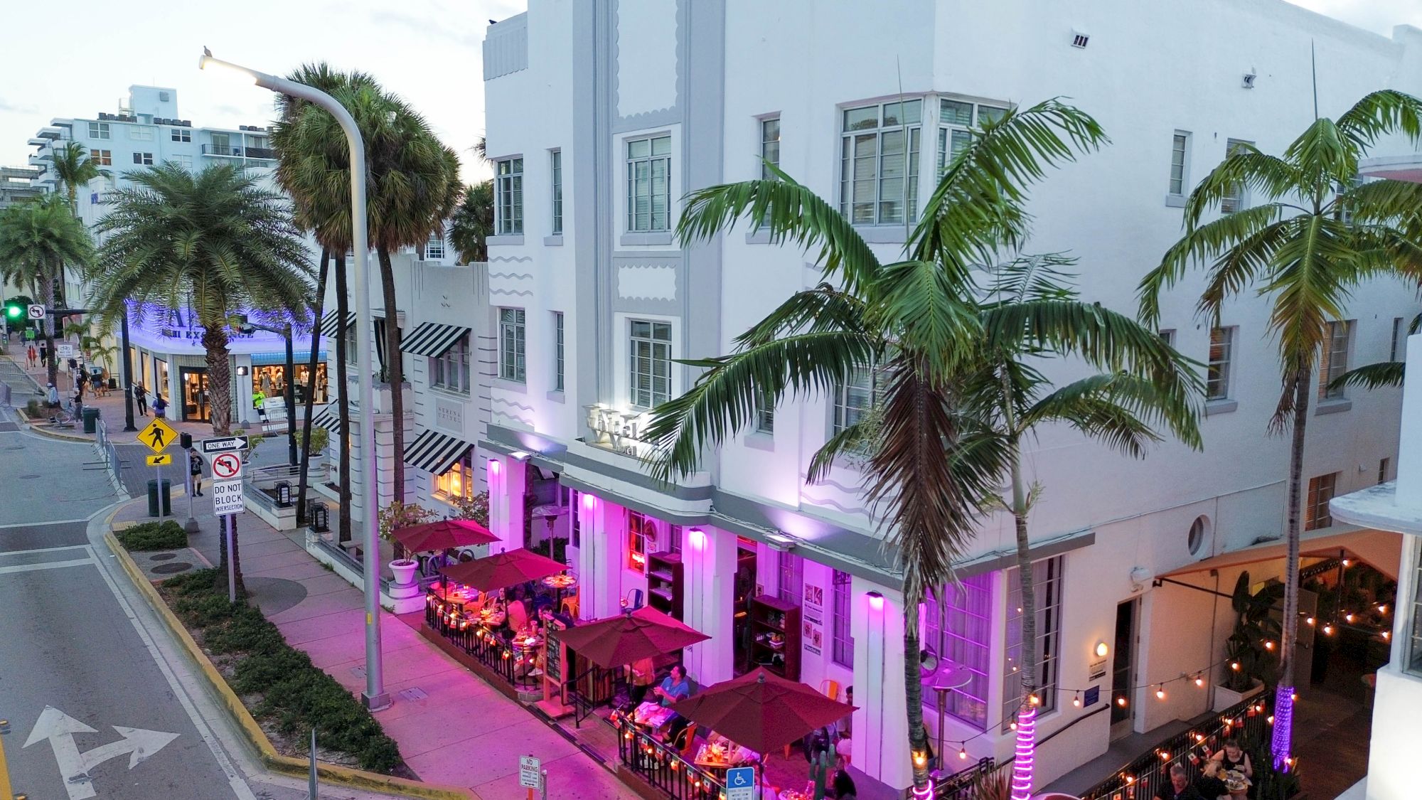 A chic street scene with a pastel-colored building, outdoor patio seating under umbrellas, palm trees, and purple lighting along a lively urban corner.