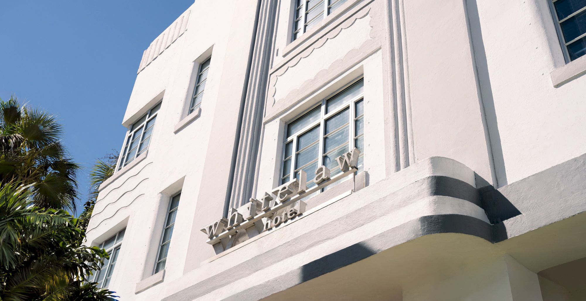 An art deco white building with rounded corners and palm trees, sign reads &ldquo;Windelay &hellip;&rdquo; or &ldquo;Lown a&hellip;&rdquo; on the fa&ccedil;ade, sunny day.