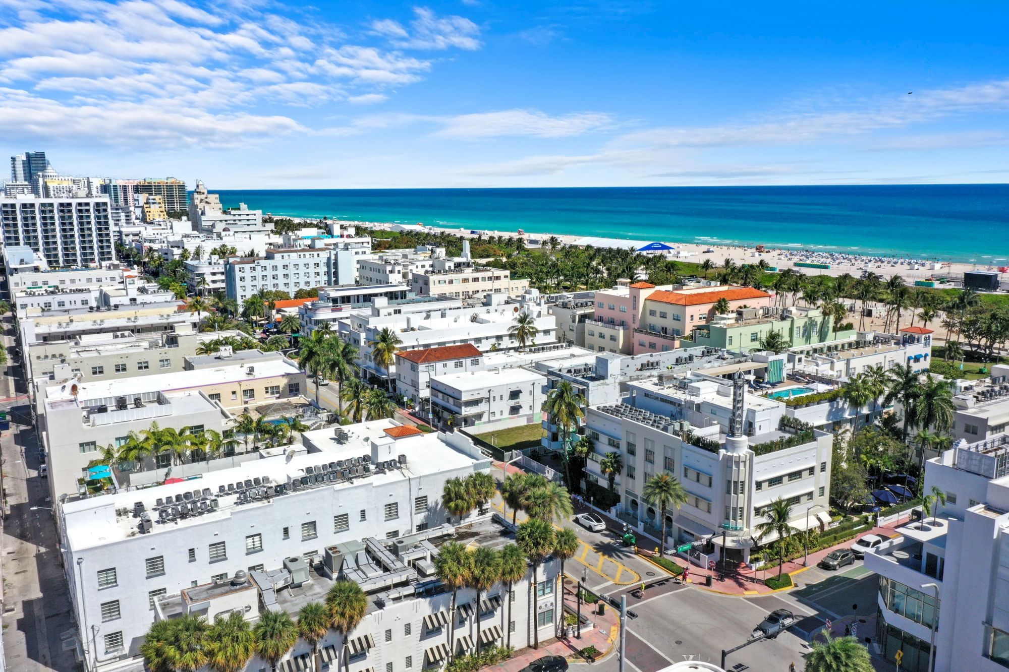 A sunny coastal cityscape with white low-rise and mid-rise buildings, palm trees, and a turquoise ocean plus a blue sky in the distance.