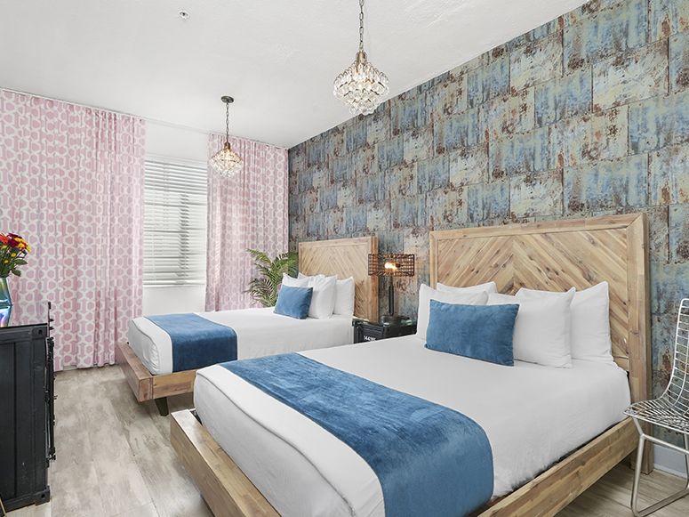 Two beds in a colorful hotel room with blue accents, patterned wallpaper, a chandelier, a TV, flowers, and a chair.