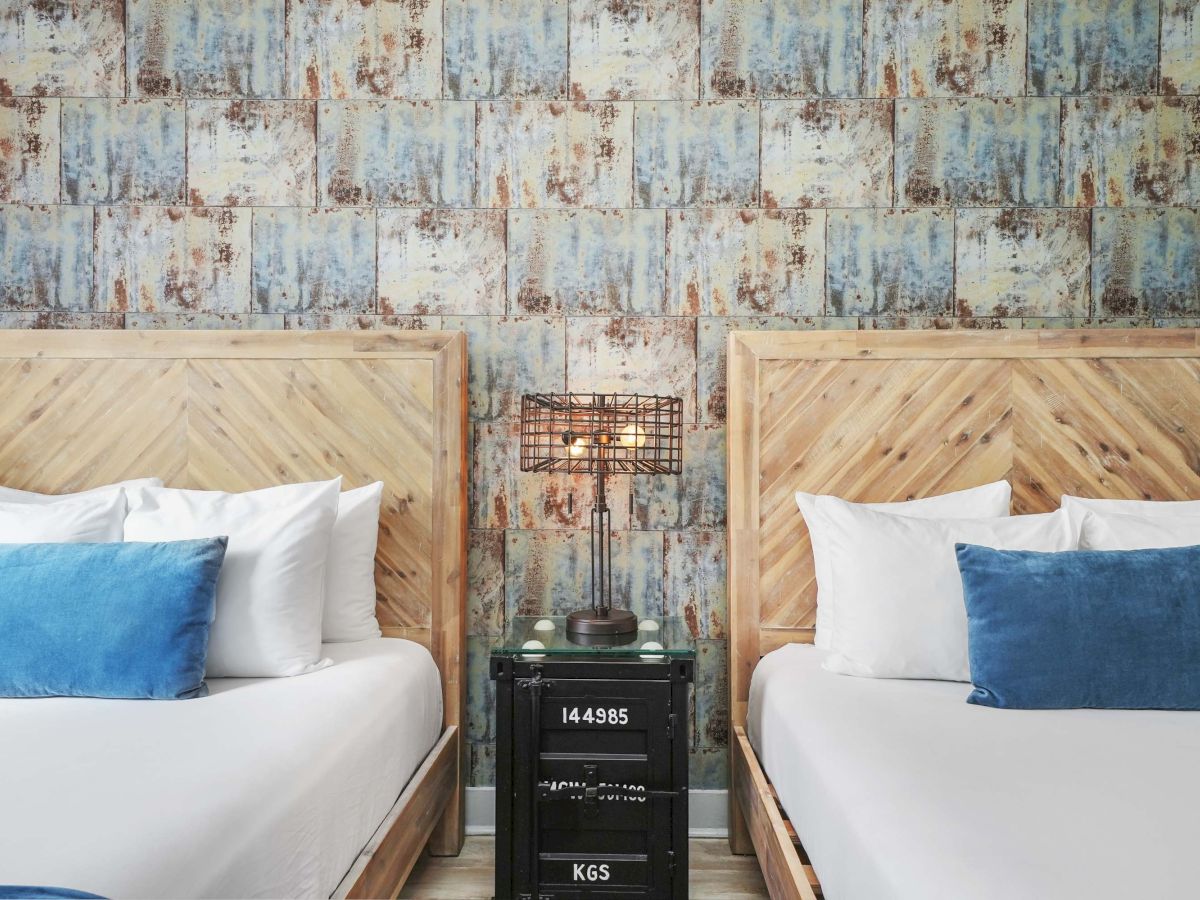 Two neatly made twin beds with matching wooden headboards and blue throw pillows, flanking a black nightstand with a small lamp, in a rustic patterned room.