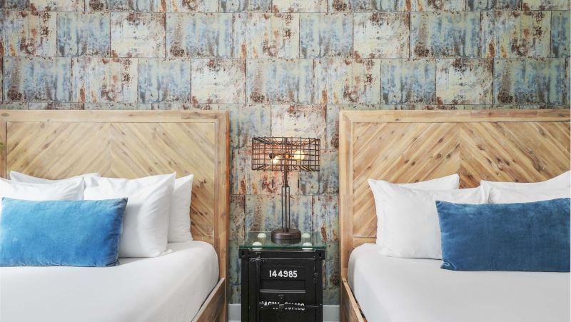 A hotel room with two wooden beds, a glass top nightstand, and an industrial style lamp.