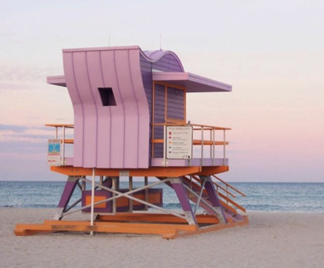 A pink lifeguard tower stands on a sandy beach, calm ocean in the background, pastel sunset sky, and wooden platform under the chair structure.