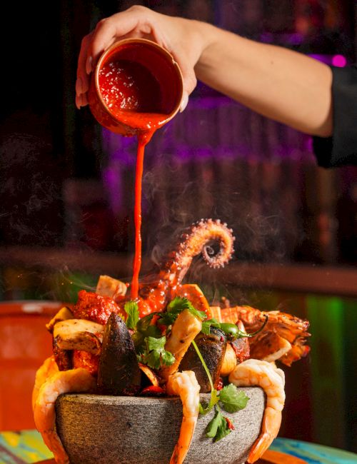 A hand pours red sauce over a spicy seafood hot pot in a stone bowl, topped with herbs and veggies for a fiery, indulgent dish.