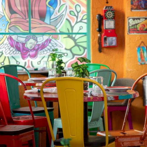 A colorful, cozy cafe with mismatched chairs around a round table, vibrant mural on the wall, plants, and warm decor.