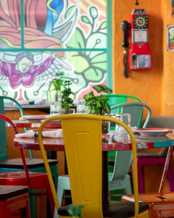A colorful, cozy cafe with mismatched chairs around a round table, vibrant mural on the wall, plants, and warm decor.