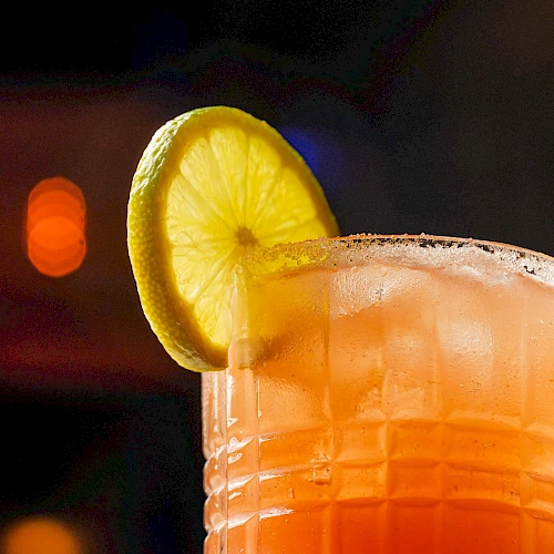 A refreshing orange-pink cocktail in a textured glass with a lemon wheel garnish on the rim, set against a dim, blurred background. It's a vibrant, upscale drink.