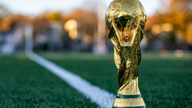 A golden FIFA World Cup trophy sits on a green football pitch near the white line, with a blurred stadium background.