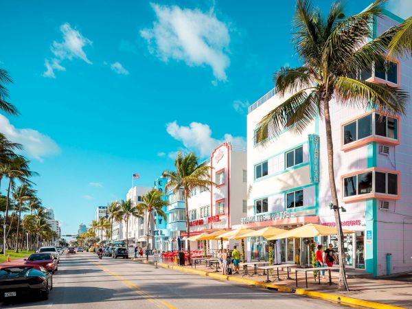 Colorful beachside street with pastel buildings, palm trees, blue sky, and pedestrians along a sunny promenade.