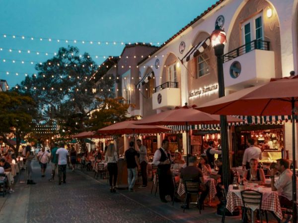 A lively outdoor street cafe scene at dusk, string lights, people dining under red umbrellas, charming buildings, relaxed evening vibe.