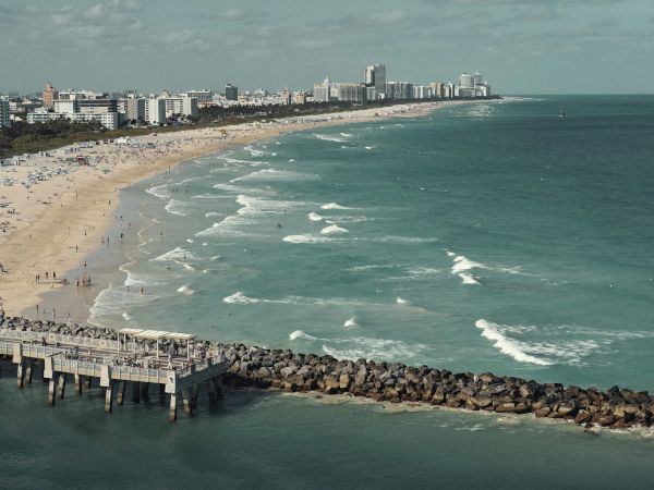 A sunny beach with a sandy shore, a rocky jetty, and calm waves; buildings line the coast in the distance.