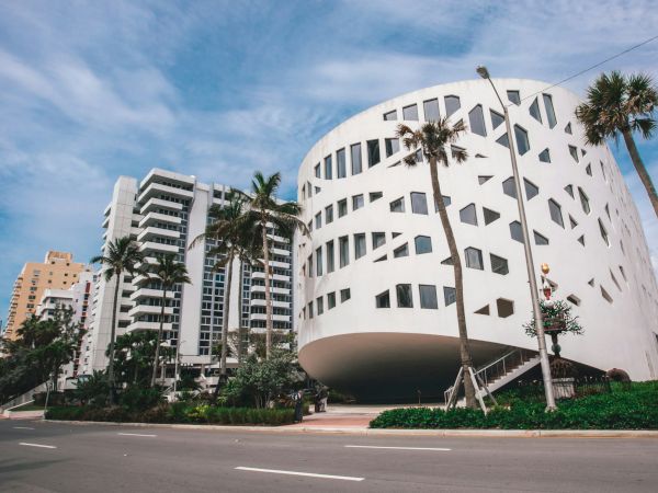 Modern white cylindrical building with rounded windows, palm trees, and a blue sky along a city street.