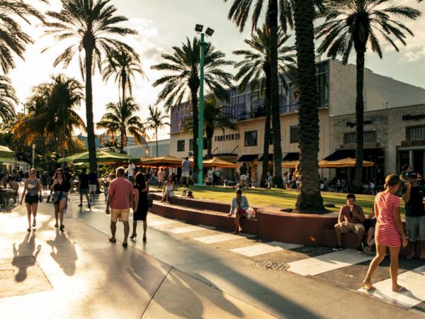 A sunny boardwalk scene with palm trees, people strolling and sitting, a shopping area in the background, and long shadows on the pavement.