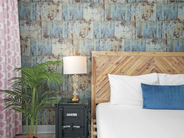 A stylish hotel-style bedroom with a wooden headboard, blue accent pillow, bedside lamp, black nightstand, plant, and pink patterned curtain.