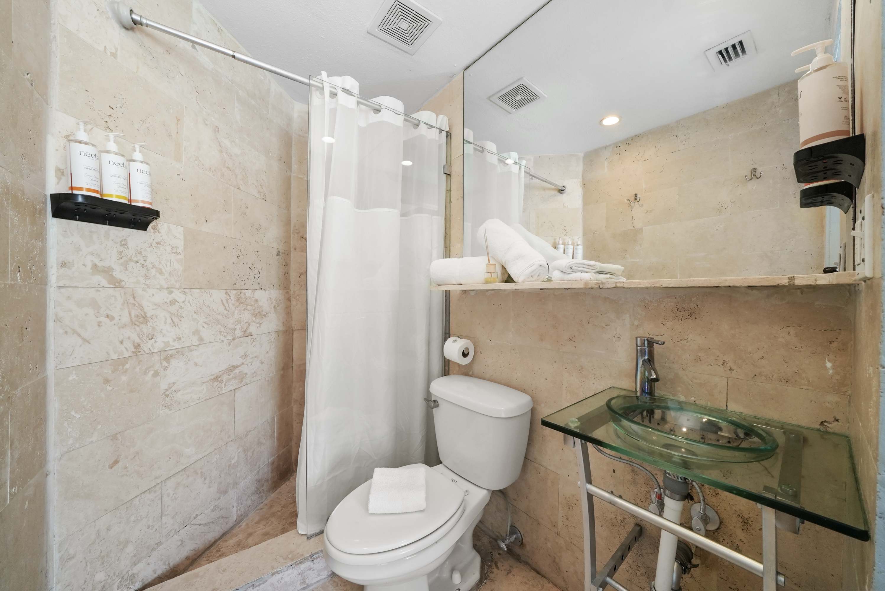 A standard hotel bathroom with a glass sink, tiled shower, toilet, and mirror.