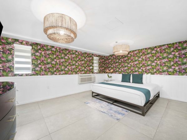 A modern bedroom with bold floral wallpaper, a white platform bed, teal accents, and minimal furniture in a bright, airy space.
