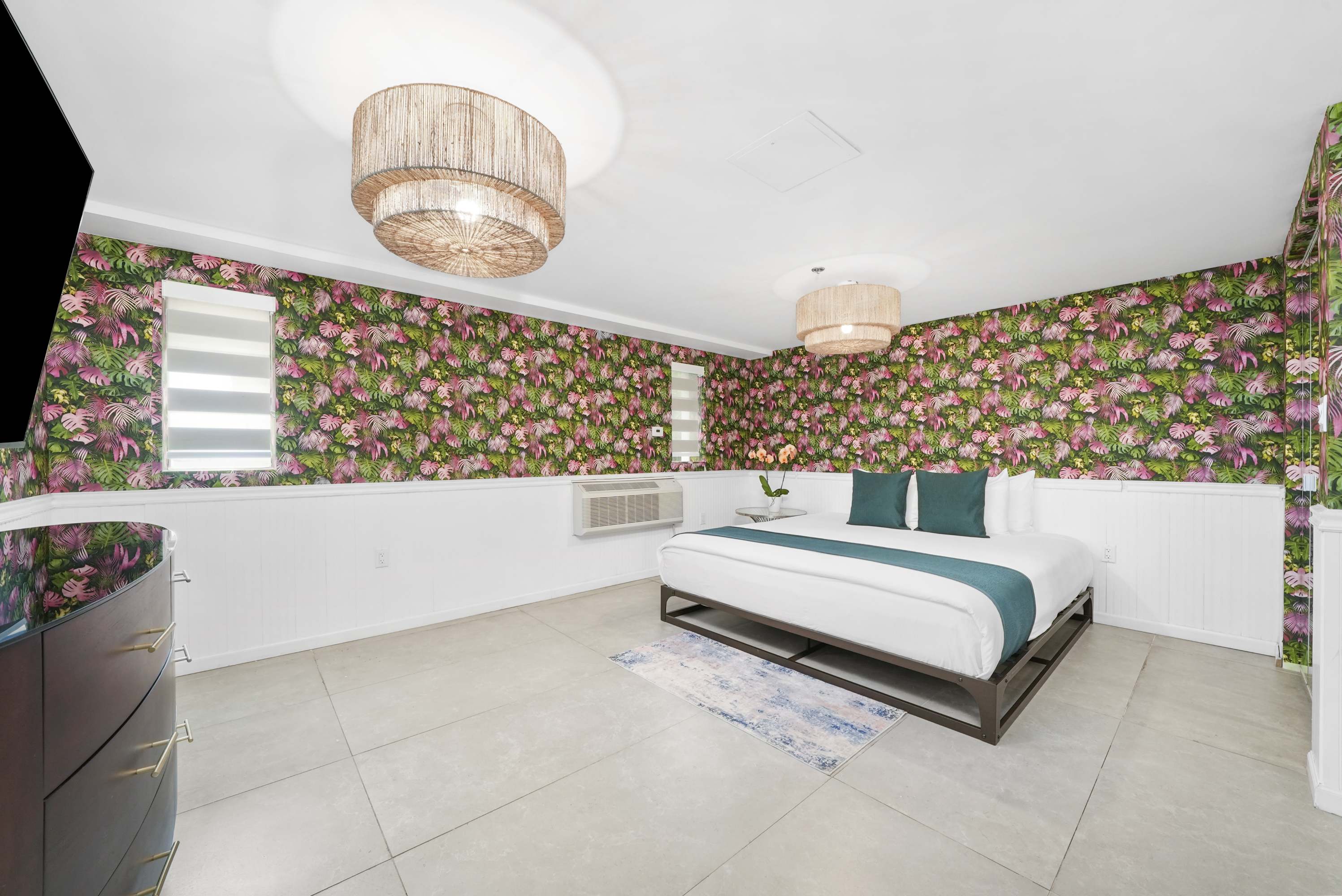 A wide-angle view of a hotel room with a floral wallpaper, a white bed, and a dark wood dresser.