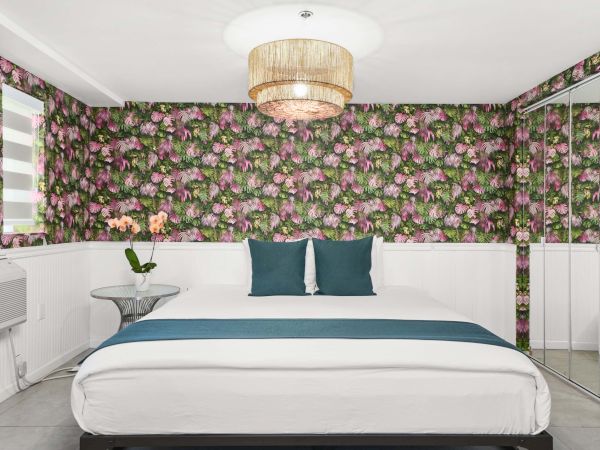 A modern bedroom with floral wallpaper, a white bed with teal accents, a round chandelier, mirrored wardrobe, and a small side table with a flower vase.