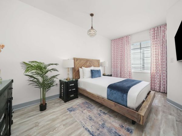 A bright, modern bedroom with a wooden bed, navy accents, pink curtains, a flat-screen TV, a dresser, plant, and soft rug, airy and tidy.