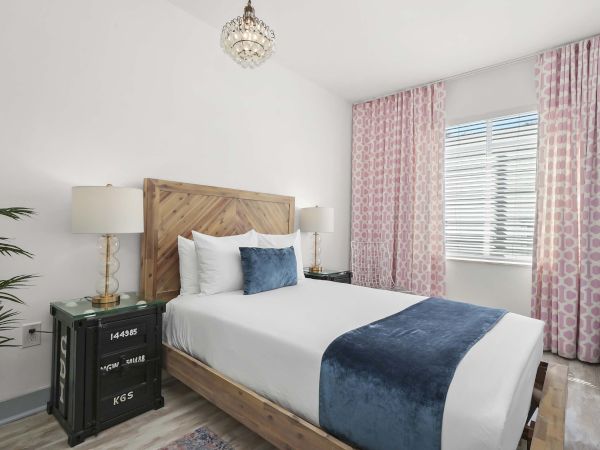A stylish bedroom with a wooden bed, white linens, navy blue throw and pillows, pink patterned curtains, two matching nightstands, and a chandelier.