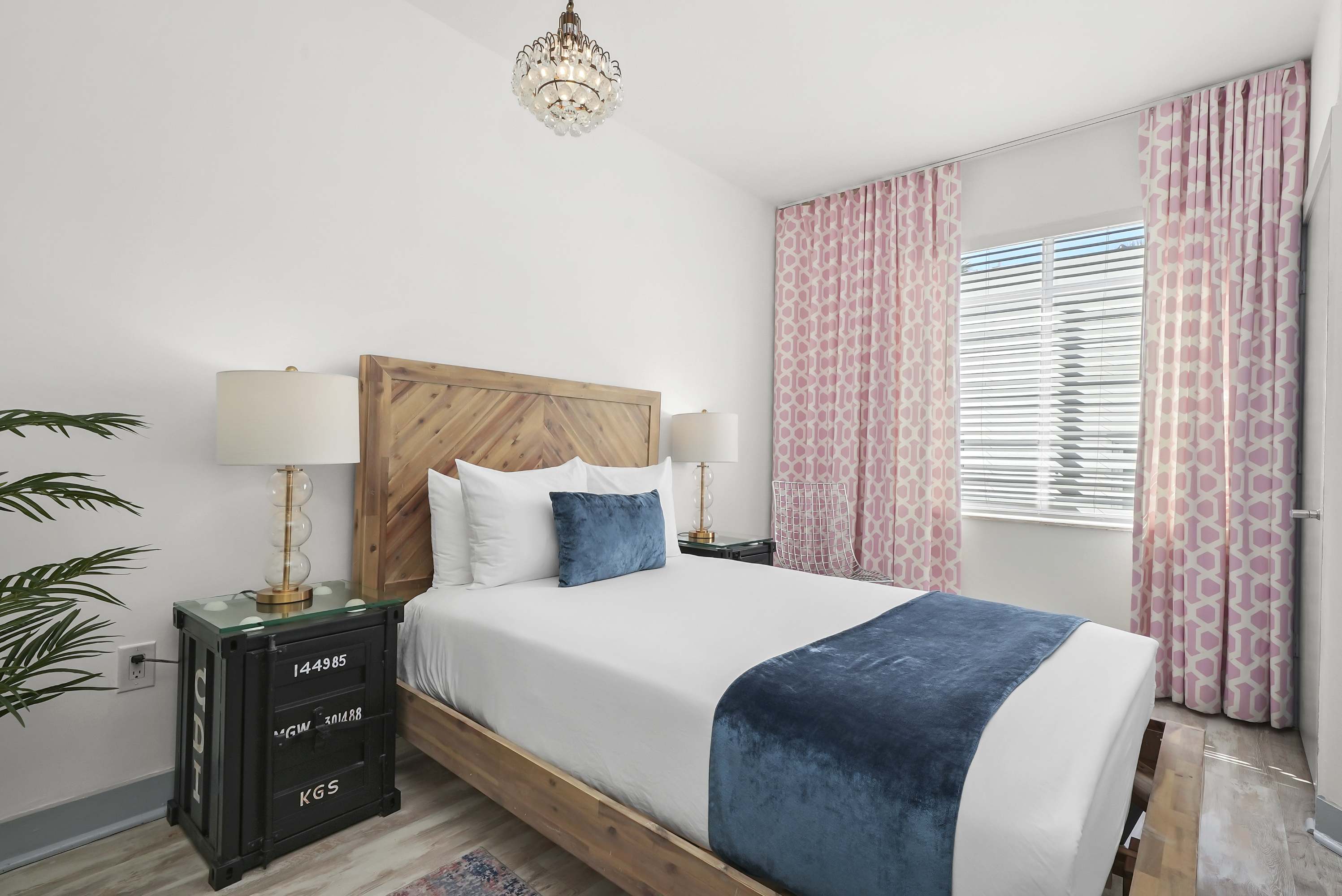 A neatly made bed with white and blue linens in a room with a window and pink curtains.