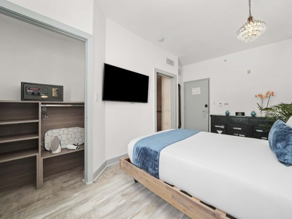 A modern bedroom with a wooden bed frame, white and blue bedding, a wall-mounted TV, and a small open closet area; bright, airy decor.