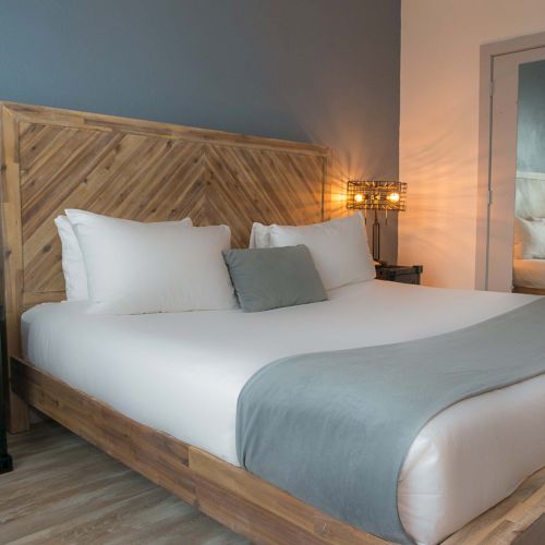 A modern hotel room with a wooden platform bed, white linens, gray throw, matching pillows, a nightstand, wall lamp, and a mirror by the doorway, glowing softly.