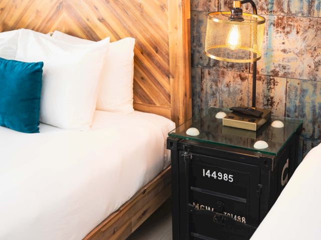 A close-up view of a bed with a wooden headboard, white bedding, and a blue accent pillow, accompanied by a unique glass-topped table with an industrial-style lamp.