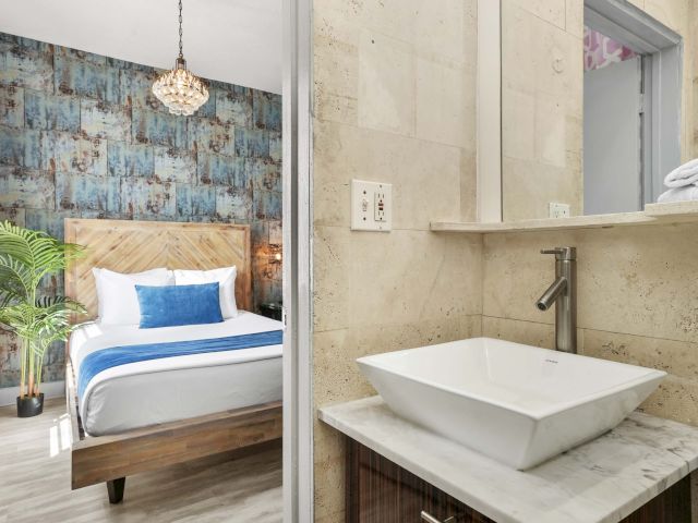 A bathroom sink area with a white sink, marble countertop, and a mirror, adjacent to a bedroom with a bed and blue accent wall.