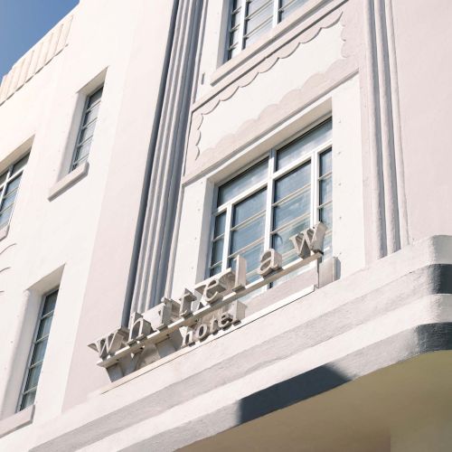 A white Art Deco building with geometric detailing and tall windows; a palm tree on the left, and a curved entrance canopy under a sign.