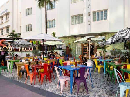 Outdoor cafe with colorful plastic chairs under umbrellas, a few people seated, and a white building backdrop.