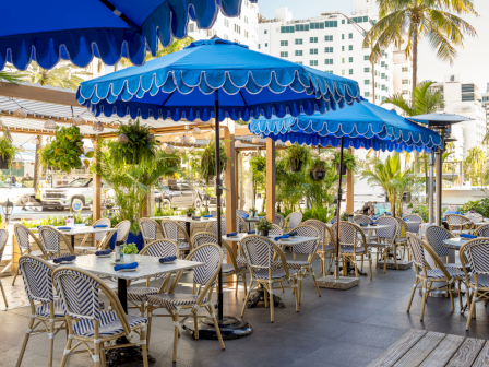 A sunny outdoor cafe with blue striped umbrellas, numerous wicker tables and chairs, potted plants, and palm trees in a lounge-like dining area.