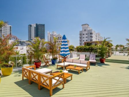 A sunny rooftop terrace with wooden lounge chairs, a blue striped umbrella, potted plants, and city buildings in the background.
