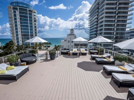 A sunny rooftop pool area with lounge chairs, umbrellas, and views of tall buildings and the ocean in the background. (140 chars)