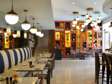 A stylish restaurant interior with a long row of tables and blue-and-white upholstered booths on the left, warm pendant lights, and a bright orange display wall with shelves and seating on the right.