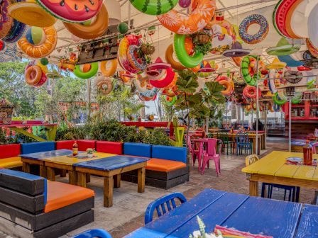 Colorful outdoor cafe with bright blue and orange seating, yellow tables, and hanging inflatable donut and pool-ride decorations. End.