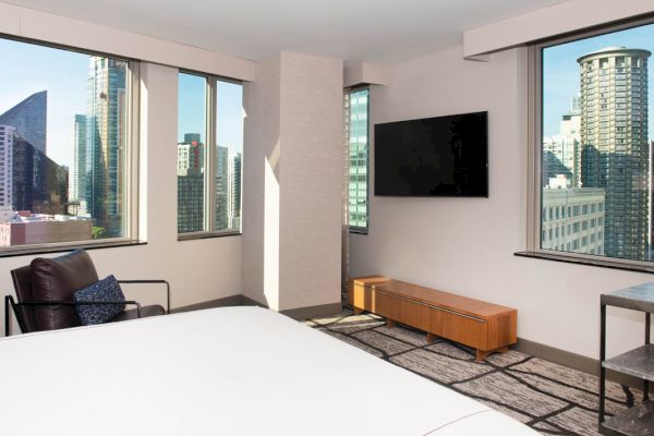 A modern hotel room with a city view, featuring a bed, chair, TV, and desk.