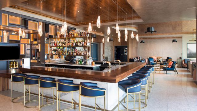 A modern bar with a long wooden counter, blue stools, and pendant lights.