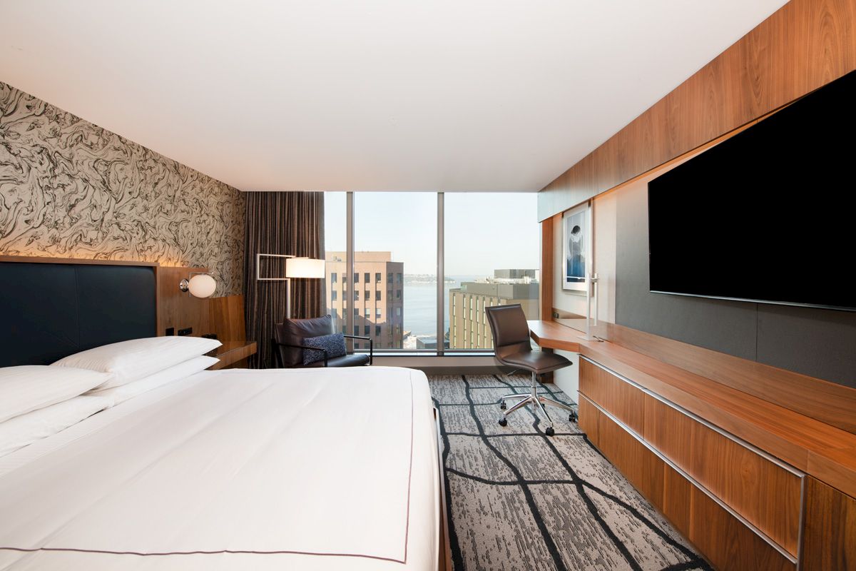 A modern hotel room with a king-size bed, desk, chair, and city view.